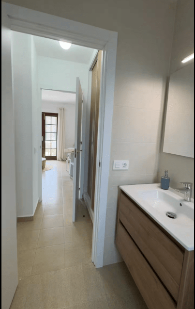 View of a modern bathroom with a wooden vanity and sink, featuring a mirror and a doorway leading to another room.