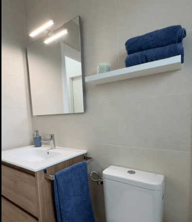 A modern bathroom featuring a white sink with a wooden cabinet, a large mirror above with stylish lighting, a shelf holding folded blue towels and a decorative item, and a white toilet beside a blue towel hanging.