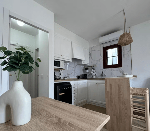 A modern kitchen featuring white cabinetry, a marble backsplash, and a small dining area with a wooden table and chairs. A potted plant is on the table, and the room has a warm and inviting atmosphere.