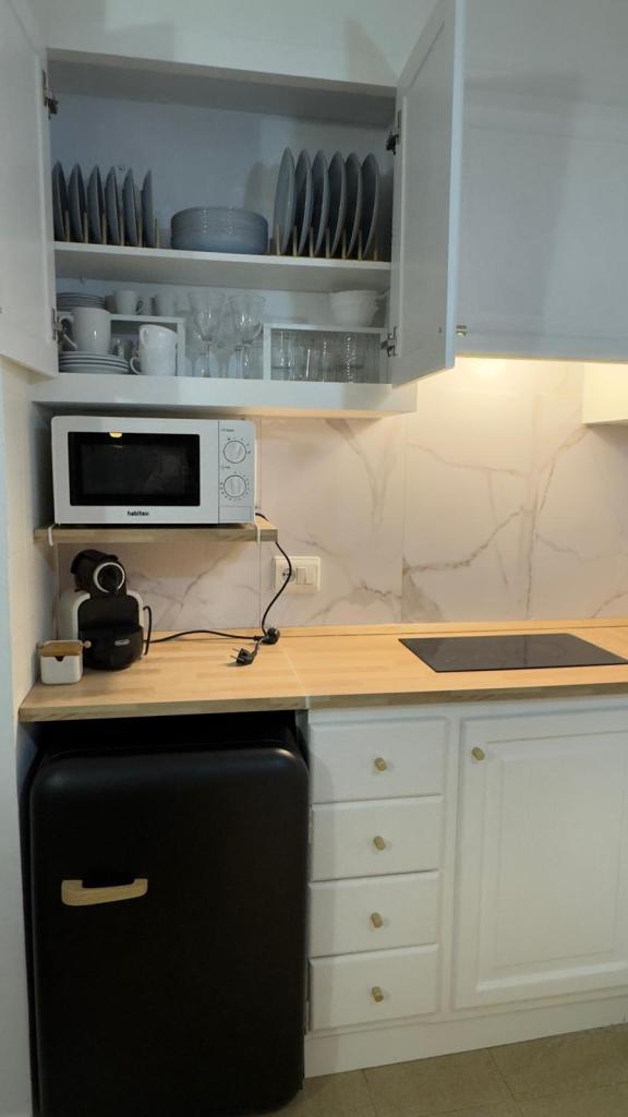 A modern kitchen corner featuring a black refrigerator, a wooden countertop with an induction cooktop, a microwave, and a coffee machine. Above the counter, there are white cabinets filled with dishes and glassware.