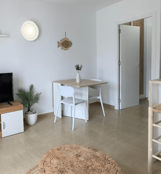 A bright and modern living space featuring a television, a small dining table with two white chairs, a potted plant, and a decorative fish wall hanging.