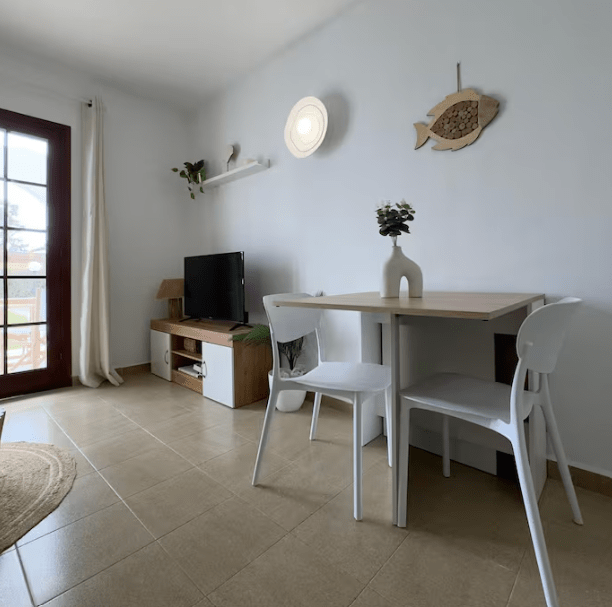 A modern living room featuring a small dining table with two white chairs, a television on a wooden stand, and decorative elements including a plant and a wall-mounted fish. Natural light enters through a window with sheer curtains.
