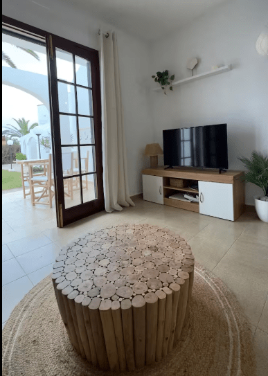 A cozy living room featuring a round wooden coffee table, a television on a wooden cabinet, and large glass doors leading to a garden. Natural light fills the space, enhancing the neutral color scheme and inviting decor.