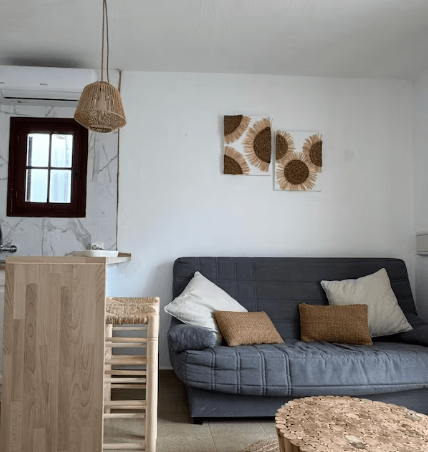 Cozy living space featuring a gray sofa with decorative pillows, a wooden bar table with stools, and wall art depicting sunflowers, set in a bright room with a window and hanging light fixture.