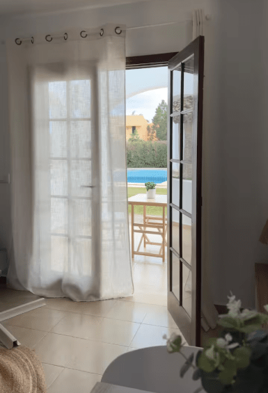 Interior view of a room with a partially open door leading to a patio and swimming pool, with sheer curtains and a small table set outside.