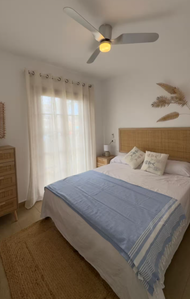 Cozy bedroom featuring a bed with a light blue throw, a rattan headboard, a wooden nightstand, and natural light streaming in through sheer curtains.