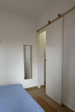 A bright bedroom featuring a blue bedspread, a small mirror on the wall, and a sliding door leading to another room.