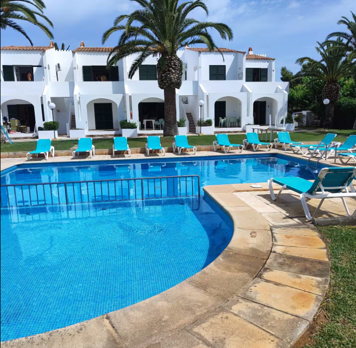 A sunny pool area featuring a blue swimming pool surrounded by lounge chairs with turquoise cushions, palm trees in the background, and white-tiled buildings.