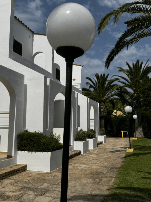 A pathway lined with round white lamps leads to a white building surrounded by palm trees and manicured bushes under a blue sky.