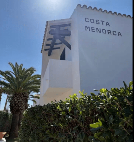 Exterior view of a building with the sign 'Costa Menorca' and palm trees in the background