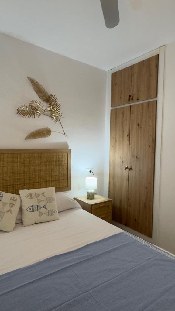 A cozy bedroom featuring a woven headboard, decorative pillows with fish patterns, a light wood nightstand with a lamp, and a wooden wardrobe. The room has a light and airy feel, accented by a ceiling fan and wall decor of dried leaves.