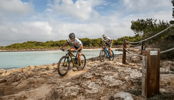 Two mountain bikers riding along a rocky path near a body of water, surrounded by greenery and a cloudy sky.