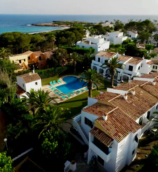 Aerial view of a coastal resort with white buildings, palm trees, and a swimming pool, overlooking the ocean.