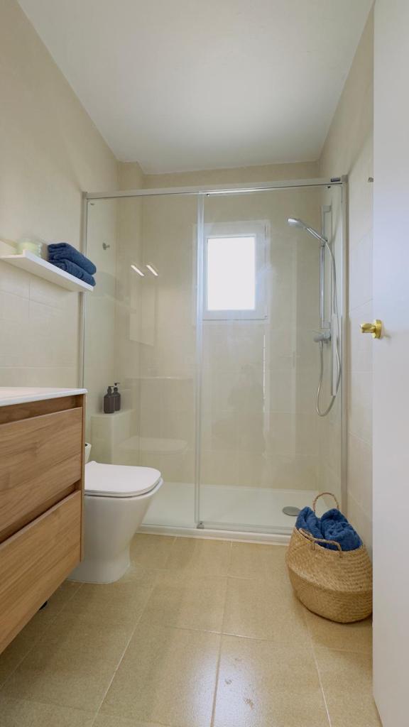 A modern bathroom featuring a glass shower enclosure, a toilet, and a wooden vanity. Towels are neatly stacked on a shelf and in a woven basket on the floor.