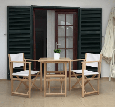 A small outdoor dining setup featuring two wooden chairs with white cushions and a wooden table, situated in front of a door with glass panels and green shuttered windows.