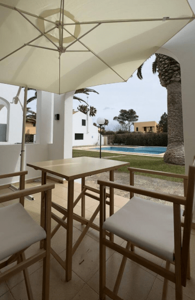 A view from a patio with wooden bar stools and an umbrella, overlooking a swimming pool and palm trees in a residential setting.